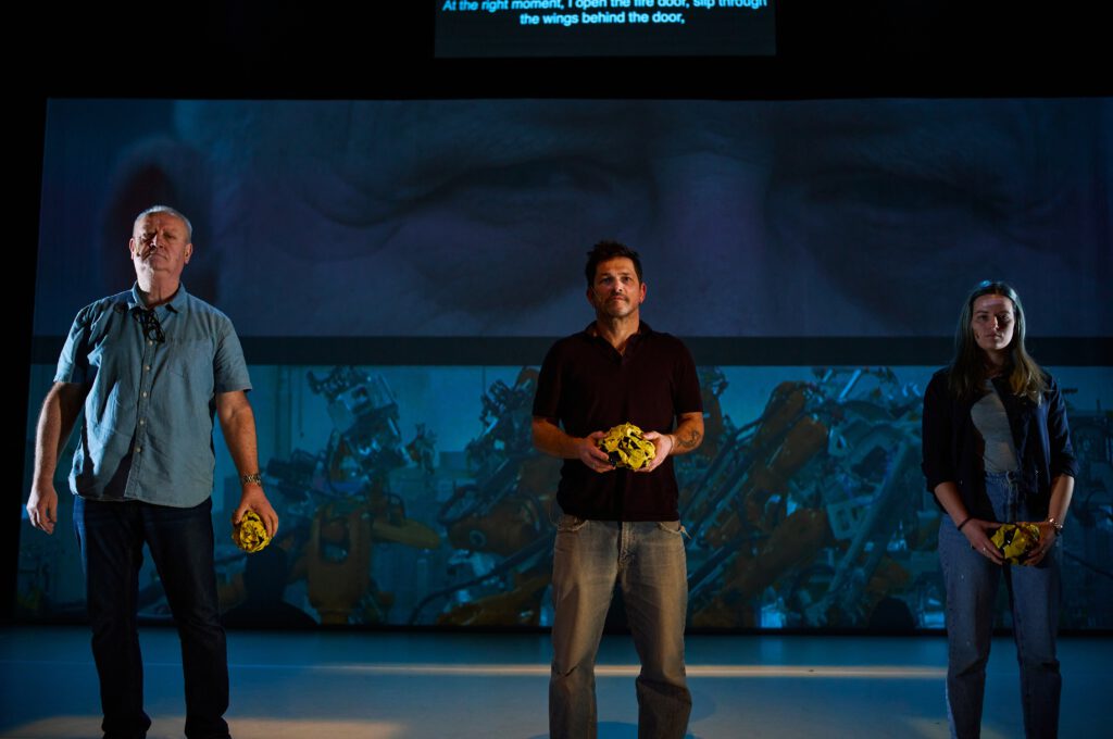 Eine dunkle Bühne, im Hintergrund eine Leinwand, auf der eine Videoprojektion zu sehen ist. Vor ihr stehen drei Personen.