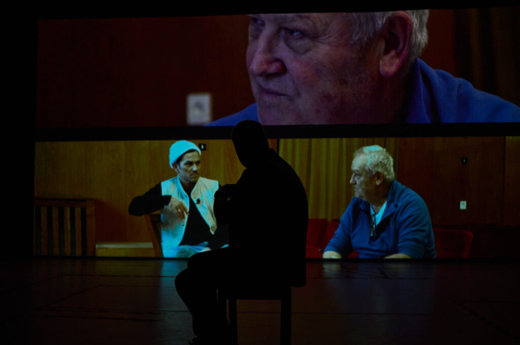 Eine dunkle Bühne, im Hintergrund eine Leinwand, auf der eine Videoprojektion zu sehen ist. Der Bildausschnitt des Videos zeigt zwei männlich gelesene Personen unterschiedlichen Alters, die sich unterhalten. Sie sitzen auf Stühlen und sind nur von ihrem Oberkörper aufwärts zu sehen. Die linke Person ist um die dreißig Jahre alt, hat dunkle Haare und trägt eine weiße Wollmütze. Über einem schwarzen Langarmshirt trägt sie eine weiße Weste, die vorne geöffnet ist. Sie scheint zu sprechen. Die rechte Person sitzt im Halb-Profil, ist um die 60 Jahre alt, hat weiße Haare und scheint zuzuhören. Sie trägt ein blaues Hemd über einem weißen T-Shirt. Die gleiche Person wird auf der Leinwand über der Konversations-Situation noch einmal in Großaufnahme gezeigt. Vor der Leinwand zeichnet sich ein schwarzer Schatten ab von einer Person, die auf der Bühne auf ihren Knien sitzt und dem Gespräch auf der Leinwand zugewandt ist.