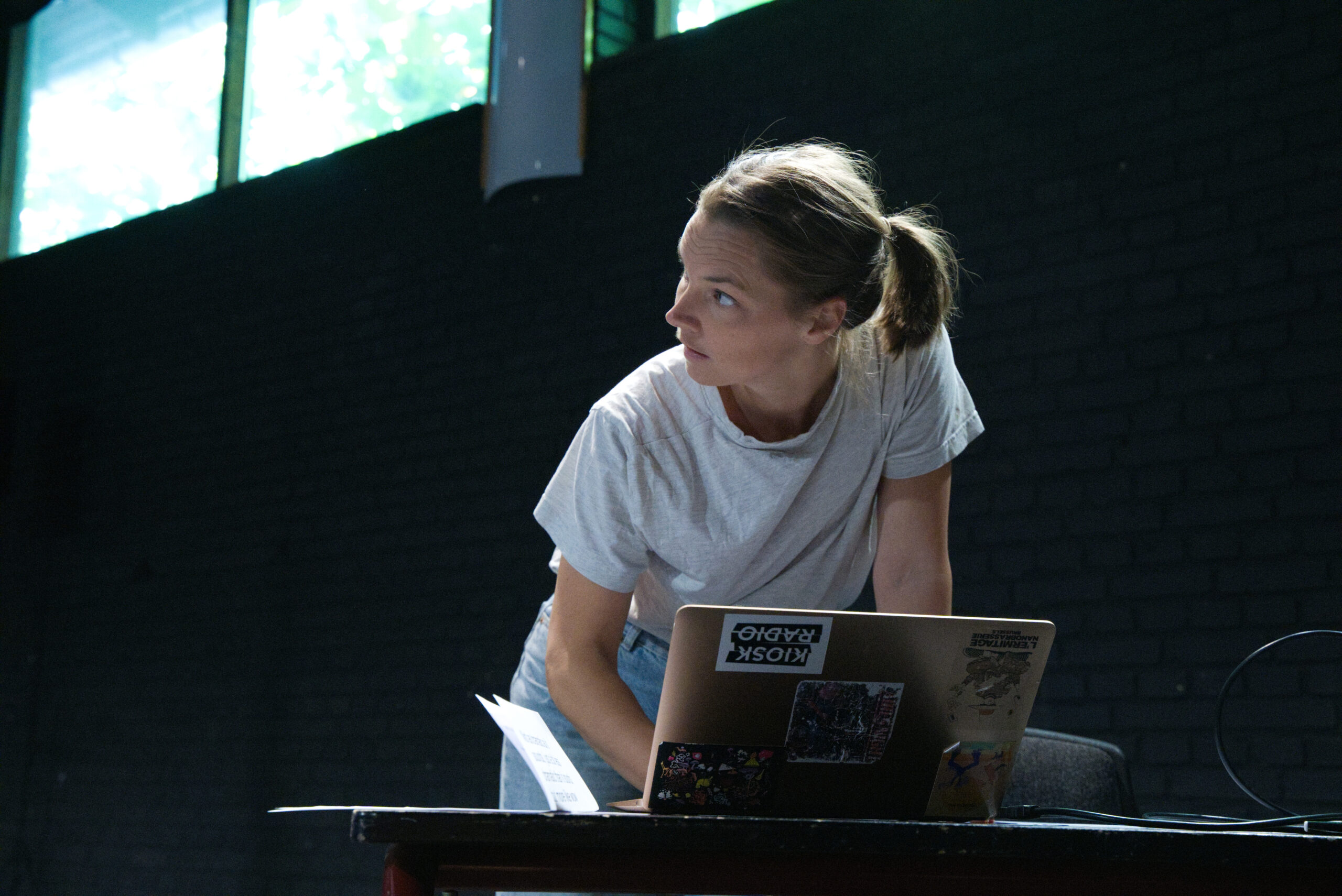Das Bild zeigt Ingrid Berger Myhre. Sie hat blonde halblange Haare, die zu einem Pferdeschwanz zusammengebunden sind. Sie trägt ein graues lockeres T-Shirt und eine Jeans. Vor ihr steht ein mit Stickern beklebter Laptop auf einem schwarzen Tisch, ein Kabel führt rechts aus dem Bild, links neben dem Laptop liegt ein Heft. Ingrid Berger Myhre bedient den Laptop mit ihrer rechten Hand, ihr Blick richtet sich aber gleichzeitig auf etwas, das links außerhalb des Bildes liegt.