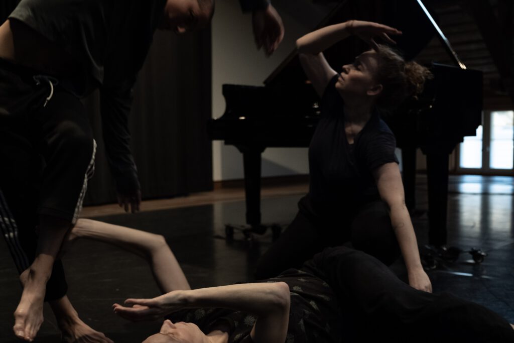 Das Bild zeigt die Darsteller*innen in einem Probenraum mit schwarzem Tanzboden. Alle sind in Bewegung, in einer körperlichen Verbindung miteinander. Im Hintergrund steht ein schwarzer Flügel.