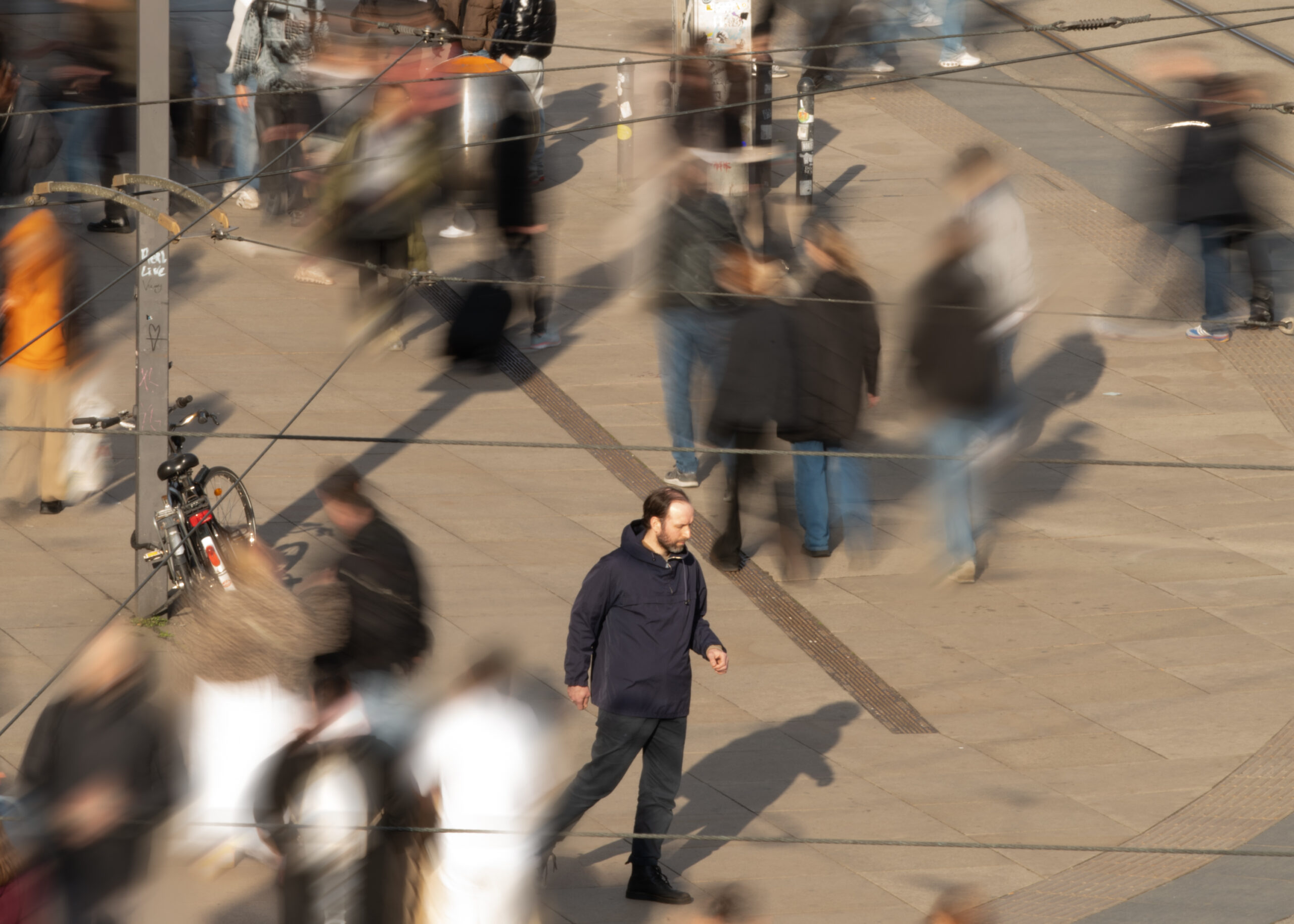 Ein großer öffentlicher Platz mit vielen Menschen, die in unterschiedliche Richtungen gehen. Durch die Aufnahme ihrer Bewegung sind sie unscharf abgebildet. Im Mittelpunkt des Bildes ist eine männlich gelesene Person mit dunkler Kleidung – einer Hose und einer funktionalen Jacke – scharf zu sehen. Er bewegt sich ebenfalls über den Platz, mit unbestimmbarem Ziel.