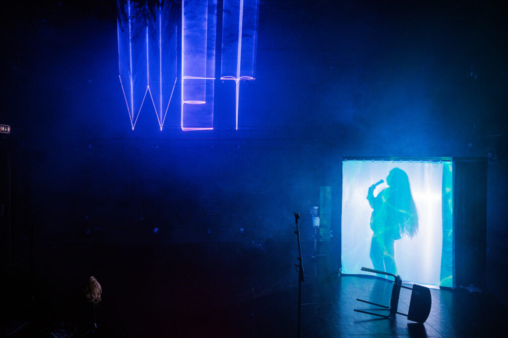 Dunkle, atmosphärisch beleuchtete Bühnenszene in Blau- und Violetttönen. Rechts steht eine halbtransparente, von innen beleuchtete Kabine oder Leinwand, hinter der die Silhouette einer Person mit langen Haaren zu sehen ist, die eine Hand vor das Gesicht hebt. Über der Bühne hängt der leuchtende Schriftzug ‚WET‘. Im Vordergrund liegt ein umgestürzter Stuhl, daneben stehen einzelne Mikrofon- oder Stativständer. Links unten ist auf einem Ständer eine Perücke oder Haarattrappe platziert. Leichter Bühnennebel verstärkt die futuristische, geheimnisvolle Stimmung der Inszenierung.