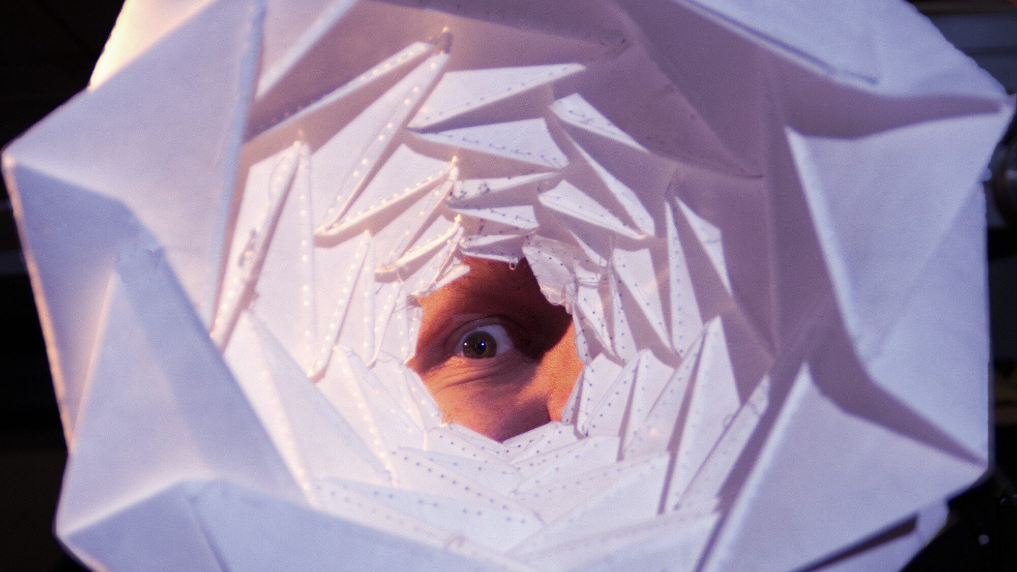 Ein Auge blickt durch einen Trichter. Wir sehen das Auge durch den Trichter hindurch. Der Trichter hat viele geometrische Falten, als sei er aus Papier gefaltet worden.