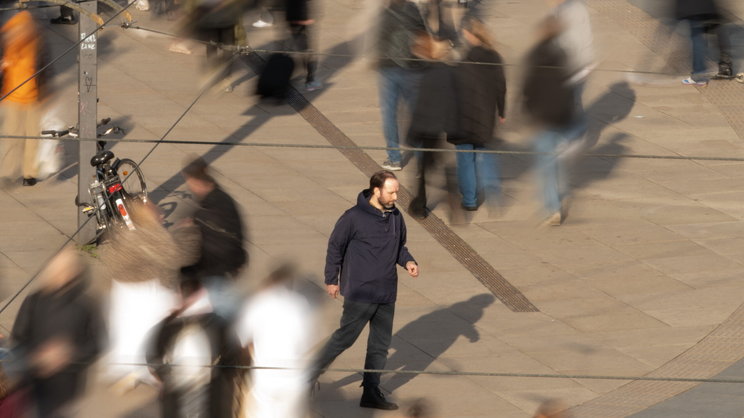Ein großer öffentlicher Platz mit vielen Menschen, die in unterschiedliche Richtungen gehen. Durch die Aufnahme ihrer Bewegung sind sie unscharf abgebildet. Im Mittelpunkt des Bildes ist eine männlich gelesene Person mit dunkler Kleidung – einer Hose und einer funktionalen Jacke – scharf zu sehen. Er bewegt sich ebenfalls über den Platz, mit unbestimmbarem Ziel.