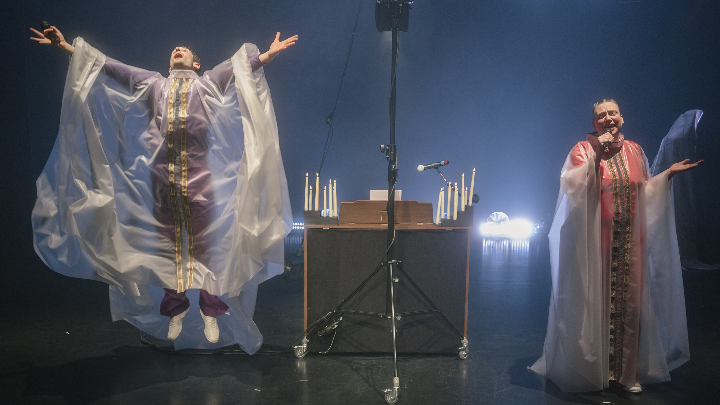 Bühnenszene in dunkler Theateratmosphäre: Zwei Personen in langen, weiten Gewändern aus halbtransparentem weißem Material mit farbigen Einsätzen stehen unter hellem Scheinwerferlicht. Links springt eine Person mit ausgebreiteten Armen in die Höhe und hält ein Mikrofon in der Hand. Rechts singt eine Person in ein Mikrofon und gestikuliert mit der freien Hand. Zwischen ihnen steht ein beleuchtetes Rednerpult oder Musikinstrument, flankiert von mehreren brennenden Kerzen. Im Vordergrund befindet sich ein großer Bühnenscheinwerfer auf einem Stativ. Die Inszenierung wirkt feierlich, musikalisch und religiös oder rituell geprägt.