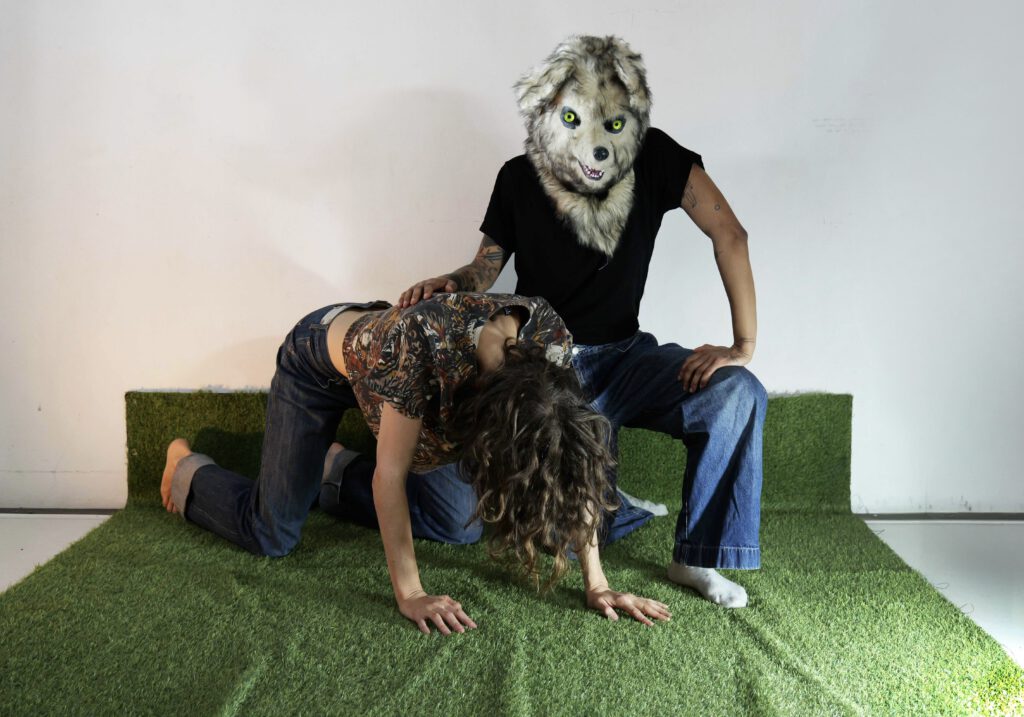 Eine Person mit einer großen, wolfsähnlichen Tiermaske sitzt auf einer grünen Kunstrasenfläche. Sie trägt ein schwarzes T-Shirt und eine weite Jeans. Vor ihr kniet eine weitere Person mit langen, lockigen Haaren, die sich mit Händen und Knien auf dem Boden abstützt. Diese Person trägt eine gemusterte Bluse und Jeans. Die Szene ist vor einer hellen Wand fotografiert.