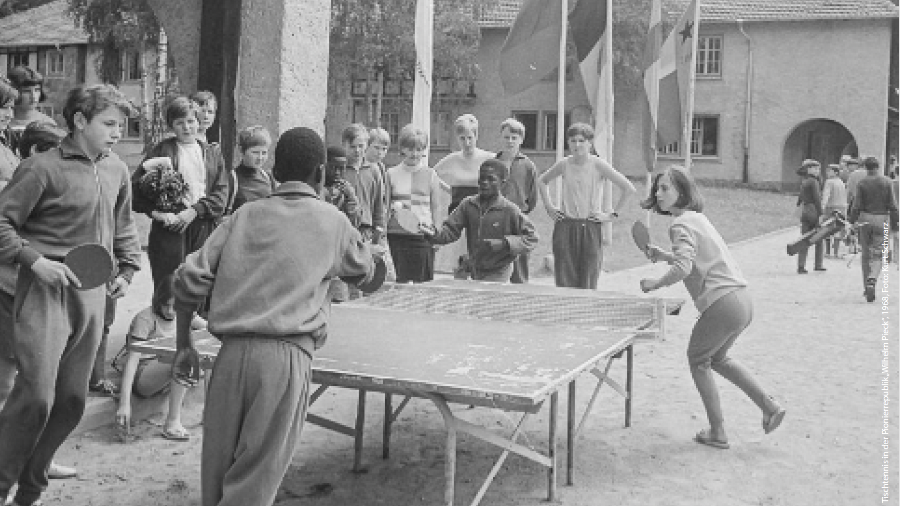 Schwarz-Weiß-Fotografie einer Gruppe von Kindern auf einem Hof zwischen Gebäuden. Im Vordergrund spielen zwei Kinder Tischtennis an einer provisorisch wirkenden Tischtennisplatte, während etwa ein Dutzend weitere Kinder das Spiel aufmerksam beobachten. Einige halten Schläger in der Hand, andere stehen in lockerer Runde um den Tisch. Im Hintergrund sind mehrere Fahnen an Masten (von denen keine konkret zu erkennen ist) sowie Gebäude mit Satteldächern zu sehen; rechts gehen weitere Kinder über den Hof. Die Szene vermittelt den Eindruck gemeinsamer Freizeitaktivitäten in einer internationalen oder gemeinschaftlichen Umgebung.