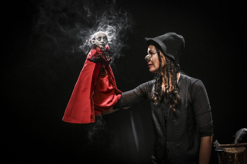 Vor komplett schwarzem Hintergrund steht eine Puppenspielerin im Profil und hält auf ihrer ausgestreckten Hand eine kleine Marionette mit weißem Gesicht, kleiner Brille und wehendem weißem Haar. Die Figur trägt einen roten Umhang mit Kapuze, um sie herum wirbelt Rauch oder Nebel. Die Spielerin trägt einen schwarzen, breitkrempigen Hut, eine kunstvolle silberne Nasenprothese in Form einer runden Kugel und lange, geflochtene dunkle Haare. Sie ist in ein schwarzes, langärmliges Gewand gekleidet und blickt der Puppe konzentriert ins Gesicht. Rechts unten im Bild sind auf einer dunklen Truhe eine Feder und eine kleine Axt oder ein Werkzeug zu erkennen.