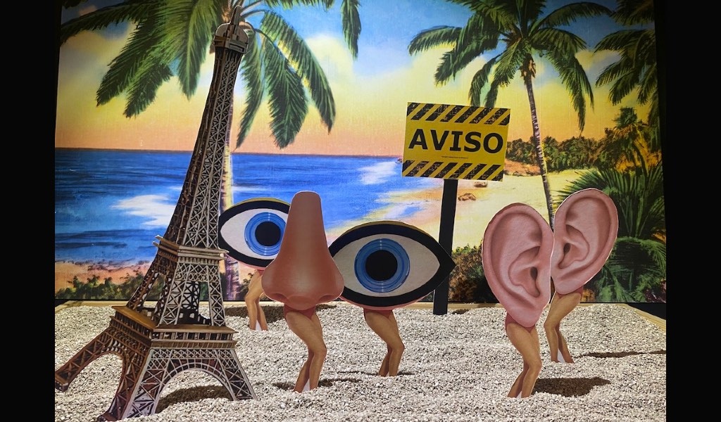 Eine surreale Miniaturszene auf einem Kiesboden vor einem bemalten Hintergrund mit Palmen, Meer und Sonnenuntergangshimmel. Links steht ein kleiner Modell-Eiffelturm. Daneben stehen mehrere absurde Figuren auf dünnen menschlichen Beinen: eine überdimensionale Nase, zwei große stilisierte Augen mit blauer Iris und ein Paar rosafarbene Ohren – alle wie kleine Kreaturen auf zierlichen Beinchen im Kies. Dazwischen ragt ein gelb-schwarzes Warnschild mit der Aufschrift „AVISO“ (spanisch/portugiesisch für „Achtung“) aus dem Boden. Die Szene wirkt wie ein skurriles Diorama, in dem Sinnesorgane als eigenständige, gehende Figuren am Strand dargestellt sind.